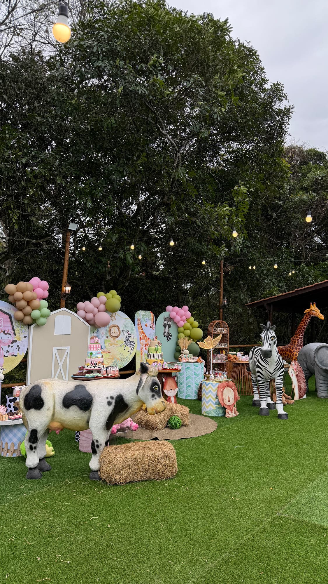 Decoração de aniversário infantil com tema de fazendinha no gramado: bichos em tamanho real, fardo de feno e arco de balões em tons pastel, sob o varal de luzes aceso