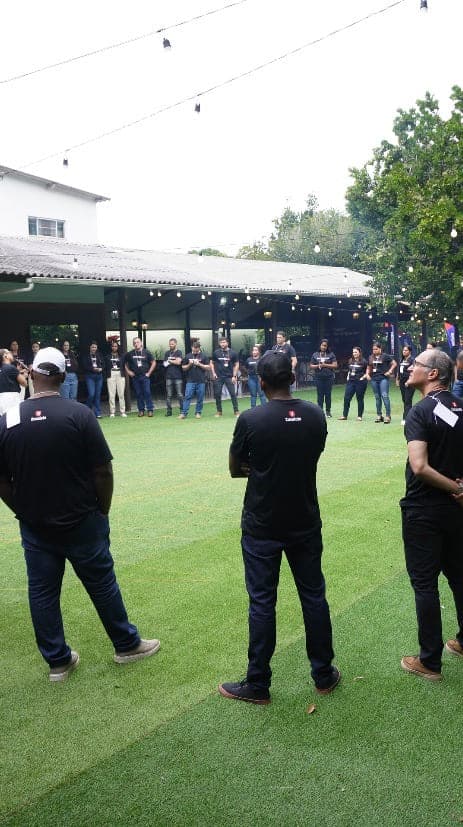Equipe de uma empresa reunida no gramado durante uma dinâmica, sob o varal de luzes, com o salão coberto e a mata ao fundo